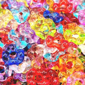 May include: A close-up shot of a pile of colorful, translucent beads. The beads are in various shapes, including circles and swirls, and in a range of colors such as red, blue, yellow, and purple. The beads are made of a shiny material.
