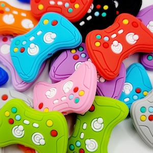 May include: A collection of colorful silicone game controller charms. The charms are shaped like a classic video game controller and feature various colors, including red, blue, green, purple, and orange.