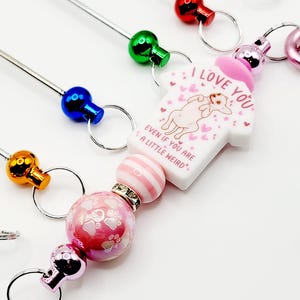 Op de afbeelding: Een roze en witte hanger met een hond die een shirt draagt met de tekst "Ik hou van je, zelfs als je een beetje raar bent" met roze hartjes en sterren. De hanger is bevestigd aan een zilveren sleutelhanger met een roze en witte gestreepte kraal en een roze kraal met witte pootjes.