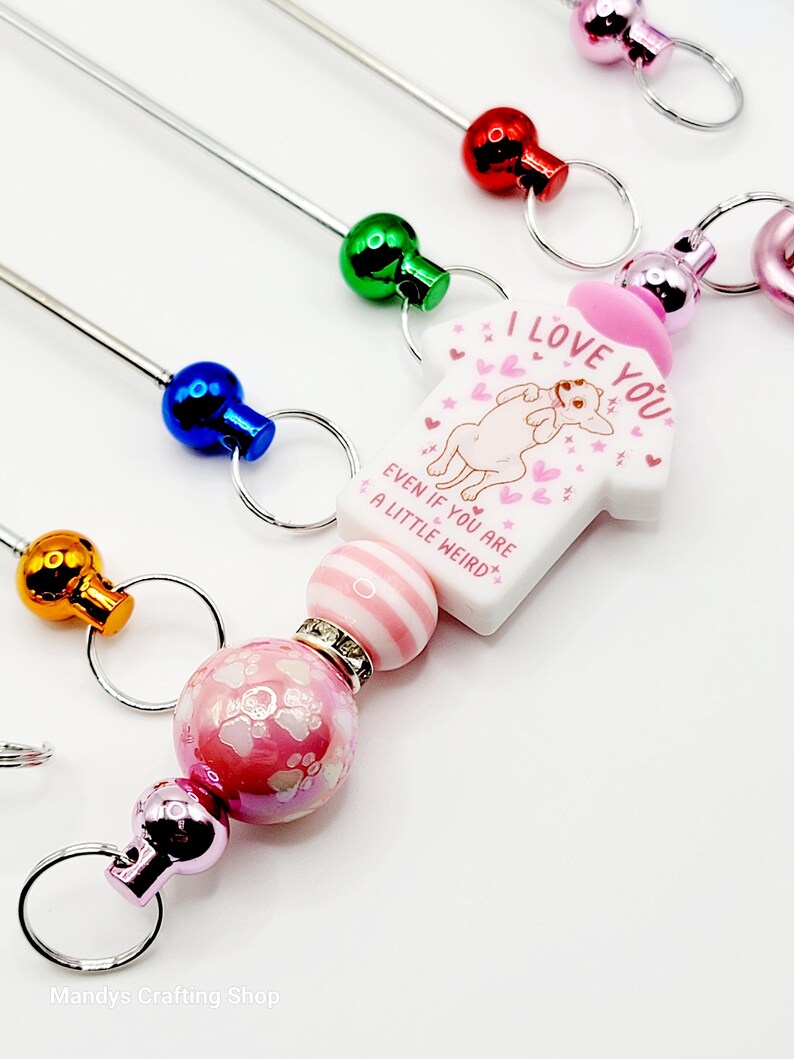 Op de afbeelding: Een roze en witte hanger met een hond die een shirt draagt met de tekst "Ik hou van je, zelfs als je een beetje raar bent" met roze hartjes en sterren. De hanger is bevestigd aan een zilveren sleutelhanger met een roze en witte gestreepte kraal en een roze kraal met witte pootjes.