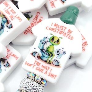 May include: A white, t-shirt shaped charm with the text "I MUST BE CONSTIPATED" and a cartoon alien on a toilet. The charm is attached to a beaded accessory with silver and iridescent accents. The text "THE WAY I DON'T GIVE A SHIT" is also visible.