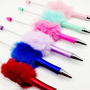 May include: A set of seven colourful pens with fluffy pom-poms on the top. The pens are in the colours white, red, blue, purple, pink, light blue, and pink. The pens are arranged in a row, with the white pen at the bottom and the pink pen at the top.