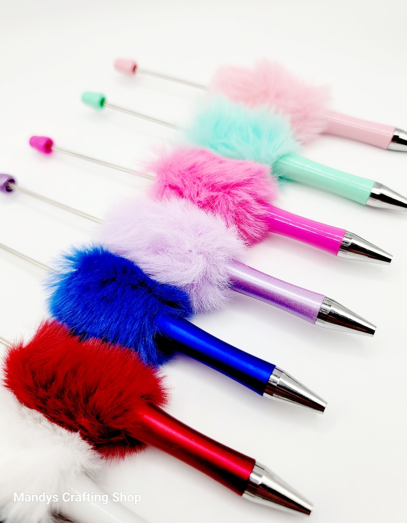 May include: A set of seven colourful pens with fluffy pom-poms on the top. The pens are in the colours white, red, blue, purple, pink, light blue, and pink. The pens are arranged in a row, with the white pen at the bottom and the pink pen at the top.