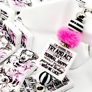 May include: A white square charm with pink and black text that reads "I'm going to try and act like a normal, happy, mentally balanced person today. Wish me luck!" The charm is attached to a pink fuzzy pom-pom and a silver and black beaded chain.