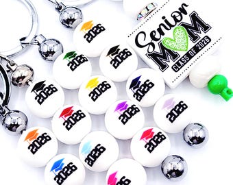 May include: A collection of graduation-themed keychains and accessories. White orbs with the year "2026" and graduation caps in various colors are featured. A rectangular charm reads "Senior MOM CLASS OF 2026". Silver and green accents complete the design.