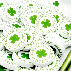 Peut inclure: Un tas de boutons ronds blancs avec un tr&egrave;fle &agrave; quatre feuilles vert et les mots "Blessed", "Lucky" et "Loved" imprim&eacute;s dessus.