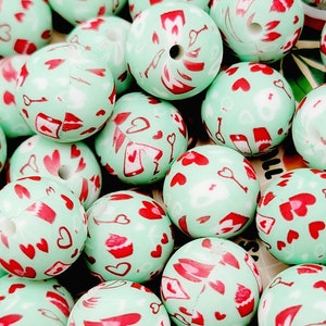 May include: A collection of mint green round beads with red heart and key designs. The beads have a hole through the center, and the surface is covered in various heart and key patterns. The beads are scattered, creating a visually appealing arrangement.
