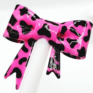 May include: A bright pink bow with a glossy finish, adorned with black spots. The bow is centered on a white cylindrical object, creating a striking contrast. The design is reminiscent of a cow print, adding a playful touch.