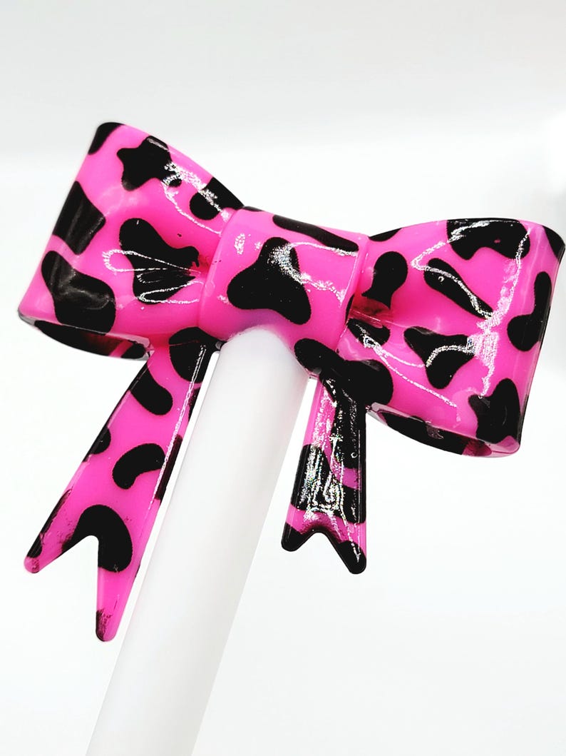 May include: A bright pink bow with a glossy finish, adorned with black spots. The bow is centered on a white cylindrical object, creating a striking contrast. The design is reminiscent of a cow print, adding a playful touch.