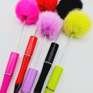May include: Five colourful pens with fluffy pom-poms on top. The pens are red, pink, purple, black, and green. The pom-poms are pink, red, black, yellow, and purple.