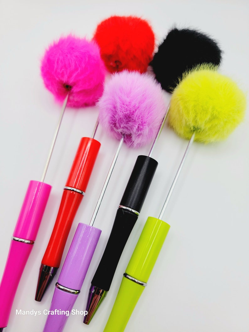 May include: Five colourful pens with fluffy pom-poms on top. The pens are red, pink, purple, black, and green. The pom-poms are pink, red, black, yellow, and purple.