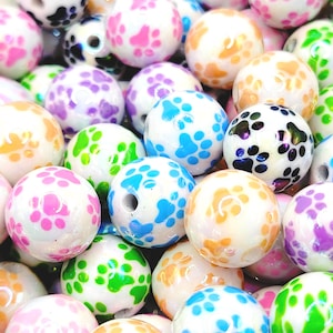 May include: A collection of colorful, round beads with paw print patterns. The beads are white with patterns in various colors, including pink, blue, green, orange, purple, and black. The beads have a glossy finish and a hole for stringing.