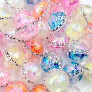 May include: A collection of round, iridescent beads featuring a central band of clear rhinestones. Each bead displays a marbled, multi-colored finish with hues of pink, blue, orange, and yellow, creating a reflective, glossy surface appearance.