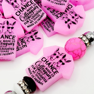 May include: Pink glitter charm shaped like a t-shirt with the text "Chance made us colleagues but the fun & laughter we share made us friends". The charm features two cartoon donkeys wearing sunglasses.