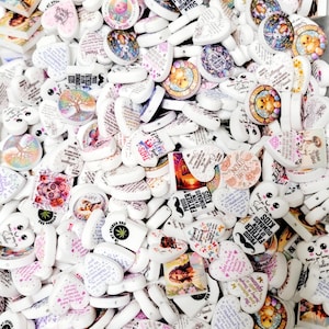 May include: A large pile of white ceramic heart-shaped and round beads with various colorful designs and text. The designs include flowers, butterflies, animals, and inspirational quotes. The beads are perfect for crafting jewelry, keychains, and other accessories.