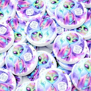 May include: A collection of round, white buttons with a watercolor design of an alien in purple and pink robes. The alien has large black eyes and is giving a peace sign. Text on the buttons reads "GREETINGS EARTH LOSER."