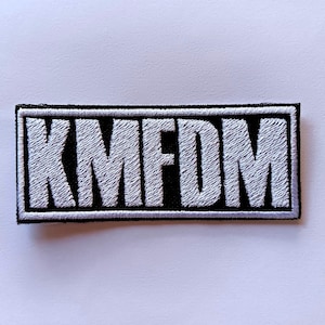 May include: Embroidered patch with the letters "KMFDM" in white, stitched onto a black rectangular background. The patch has a black border and is likely designed to be sewn or ironed onto clothing or accessories.