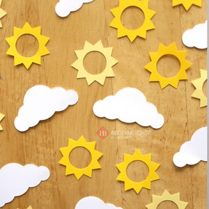 May include: A wooden surface with yellow paper cutouts of suns and white paper cutouts of clouds. The sun cutouts have a circle in the center. The clouds are simple shapes with rounded edges.