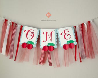 May include: A red and white tulle banner with the word "ONE" in white letters on blue and white scalloped circles. The banner is decorated with red cherries and green leaves.