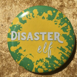 May include: A round button with a green and yellow splatter paint design. The text "DISASTER elf" is written in white on the button.