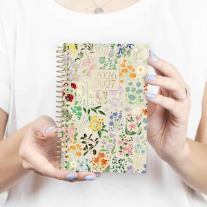 May include: A floral-patterned planner for 2026 and 2027, held by a person. The cover features a cream background with a vibrant array of colorful flowers, including red, orange, blue, and purple blooms. The spiral binding is gold.