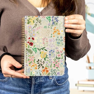 May include: A floral-patterned 2026-2027 planner with a gold spiral binding. The cover features a cream background with colorful flowers in shades of pink, yellow, blue, and orange. The planner is held by a person wearing a brown sweater and blue jeans.