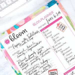 Könnte beinhalten: Ein rosa-weißer Blog-Planer mit dem Titel "bloom" und den Worten "Flying with Toddlers, Santa Fe Trip". Der Planer enthält eine Checkliste für Blog-Post-Aufgaben, darunter Entwurf, Visuals, Korrekturlesen, Rechtschreibung, Links, Formatierung, Hinzufügen von Tags, Planen/Posten, Teilen, Facebook, Instagram, Pinterest, Twitter und E-Mail. Der Planer hat auch einen Bereich für Notizen/Antworten und einen Bereich für den besten Re-Post yet! mit einem Smiley-Emoji.
