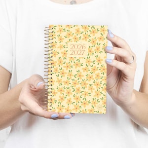 May include: A spiral-bound planner with a yellow floral pattern. The cover features small orange flowers with green leaves on a light yellow background. The years "2026" and "2027" are printed on the cover. The planner is held open.