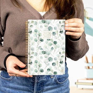 May include: A spiral-bound planner with a gold coil and a cover featuring a watercolor eucalyptus leaf design. The cover displays the years 2026 and 2027. The planner is held by a person wearing a brown sweater and blue jeans.