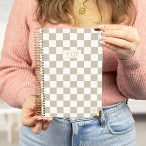May include: A spiral-bound planner featuring a beige and white checkered pattern on the cover. The center of the cover displays the year 2027 in a rectangular box. The planner includes gold-toned metal corner protectors and a gold spiral binding.