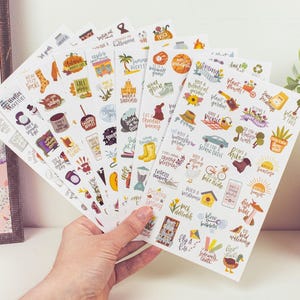 May include: A hand holds several sheets of colorful stickers with various illustrations and text. The stickers feature seasonal themes, including fall, spring, and summer activities, with phrases like "plant a tree" and "go for a bike ride."