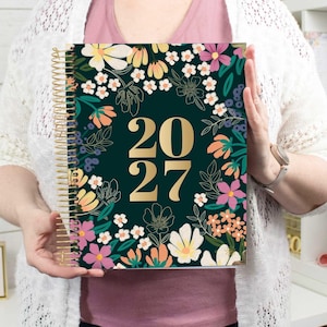 May include: A 2027 spiral-bound planner featuring a dark green cover decorated with a colourful floral pattern of pink, yellow, white, and orange flowers and green leaves. The year 2027 is printed in large, bold, gold-coloured numbers in the centre.