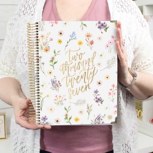 May include: A spiral-bound planner with a white cover featuring a colourful floral pattern and the gold script text "two thousand twenty seven" centred on the front. The planner has gold-toned metal spiral binding and gold-coloured corner protectors.