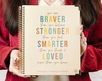 Op de afbeelding: Een beige planner met een gouden spiraalbinding. De omslag heeft een motiverende quote in kleurrijke letters: "Je bent BRAVER dan je denkt STERKER dan je voelt SLIMMER dan je denkt & GEliefd meer dan je weet." De tekst "CHRONIC ILLNESS PLANNER" staat onder de quote.