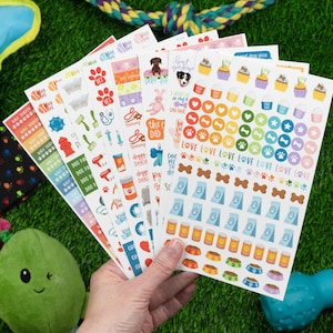 May include: A hand holds several sticker sheets with dog-themed designs. The stickers feature paw prints, dog food, cupcakes, and phrases like "Dog Days" and "Love." The sheets are on a green surface with dog toys.