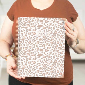 May include: A spiral-bound planner featuring a white and tan leopard print pattern across the cover. The center of the cover displays a rectangular label with the year 2027 printed in a simple, dark font. The binding is a metallic gold wire coil.