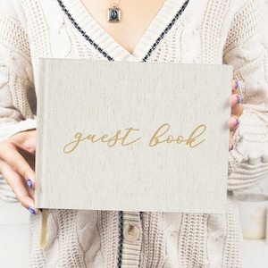 May include: A white linen guest book with gold lettering that reads "guest book".