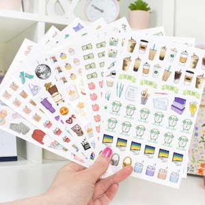 May include: A hand holds several sheets of planner stickers with various colorful illustrations. The stickers feature images of coffee cups, money, alarm clocks, and other decorative elements. The stickers are on white paper.