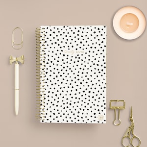 Puede incluir: Un cuaderno de espiral con cubierta blanca, estampado de manchas negras y el texto twenty seven. Los accesorios incluyen un bolígrafo blanco con un lazo dorado, un clip dorado, una pinza de encuadernación dorada y unas tijeras doradas.