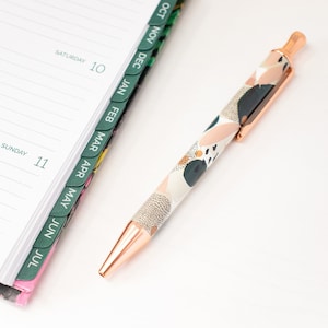 May include: A rose gold pen with an abstract pattern in shades of white, pink, and green. The pen is lying on a white surface next to a planner with monthly tabs.
