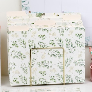 May include: File folders with a botanical leaf pattern, held in a gold-coloured metal stand. The folders are white with green leaves and small white flowers. A calendar and books are in the background.