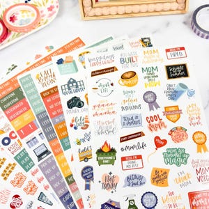 May include: A collection of adhesive sticker sheets featuring various colorful illustrations and text phrases related to parenting, school events, and daily life, including labels like Mom Life, Field Day, Pizza Party, and various motivational quotes.