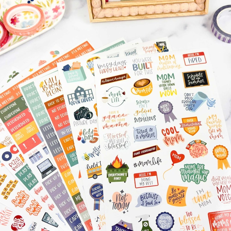 May include: A collection of adhesive sticker sheets featuring various colorful illustrations and text phrases related to parenting, school events, and daily life, including labels like Mom Life, Field Day, Pizza Party, and various motivational quotes.