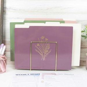 May include: A set of file folders in shades of purple, green, and pink, held in a gold-colored metal holder. The front folder is purple with a gold floral design. Papers with text are visible in the foreground.