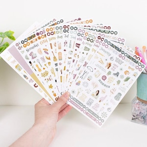 Puede incluir: Una mano sosteniendo una colección de hojas de pegatinas con varios diseños y texto. Las pegatinas presentan temas de temporada, incluyendo texto como "Noviembre", "Enero" y "Feliz Año Nuevo". Las pegatinas son de varios colores.