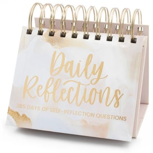 May include: A white and gold desktop calendar with a spiral binding. The front cover features the text "Daily Reflections" and "365 Days of Self-Reflection Questions".