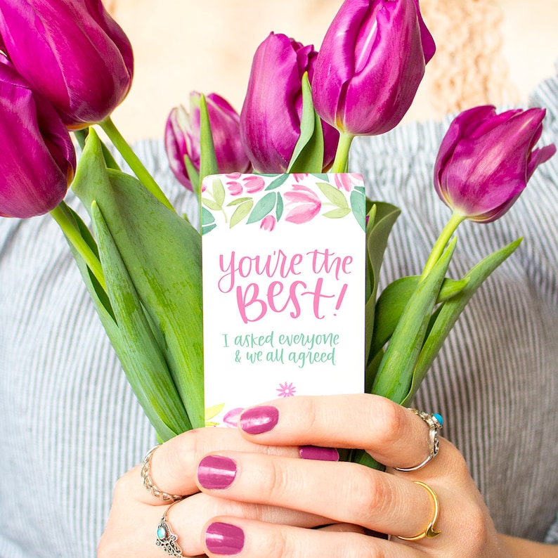 May include: A pink and green card with the text "You're the best! I asked everyone & we all agreed." The card is held by a person with pink nail polish and a bouquet of purple tulips.