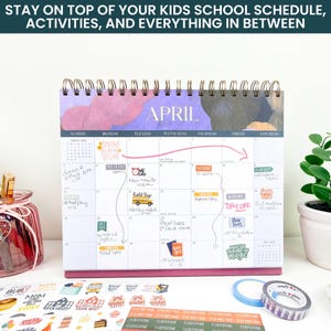 May include: A spiral-bound desk calendar for April with a purple and dark gray abstract header. The grid features handwritten notes, colorful stickers, and a pink arrow. Below the calendar are sheets of school-themed stickers and two rolls of tape.