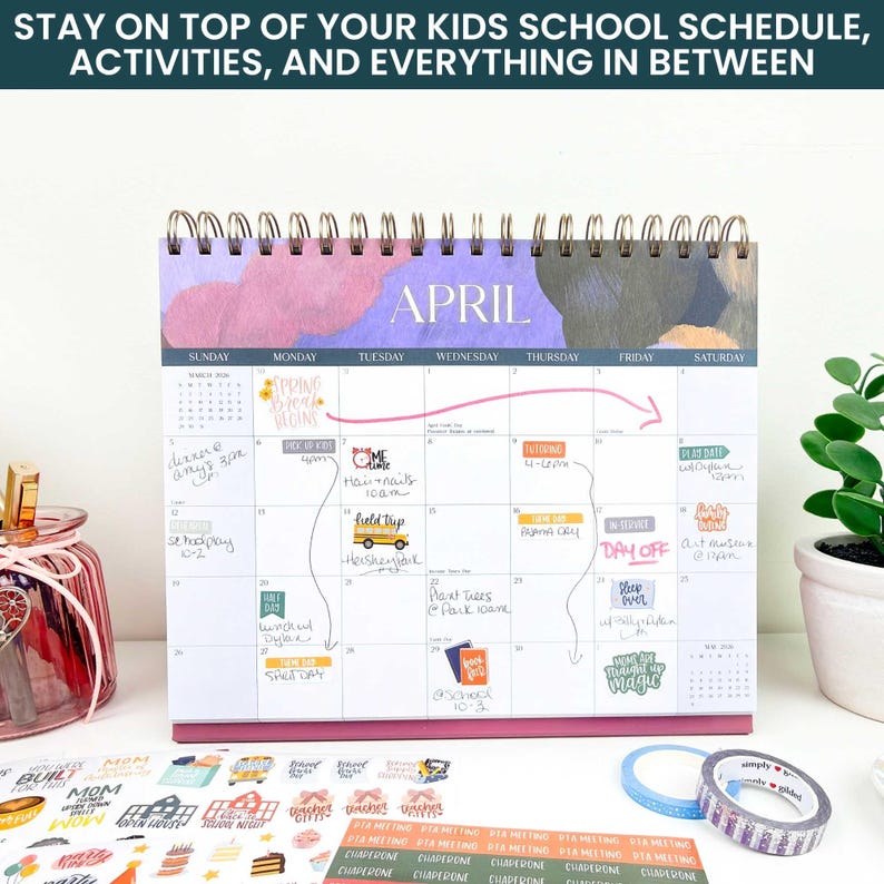 May include: A spiral-bound desk calendar for April with a purple and dark gray abstract header. The grid features handwritten notes, colorful stickers, and a pink arrow. Below the calendar are sheets of school-themed stickers and two rolls of tape.