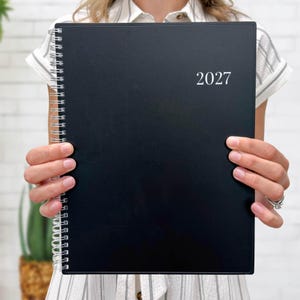 May include: A black spiral-bound planner with the year 2027 printed in white text on the front cover. The planner features a vertical silver metal coil binding along the left side and a solid matte black finish on the rectangular front cover.